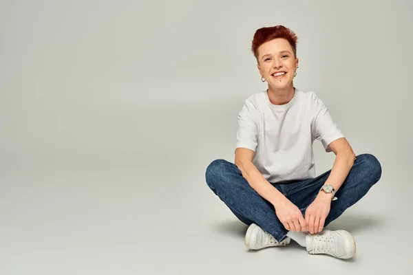 cheerful redhead non-binary person in white t-shirt and jeans sitting and looking at camera on grey