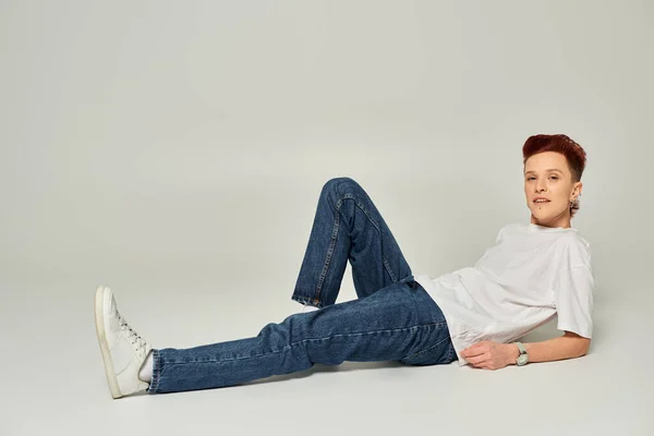 redhead queer person in white t-shirt and jeans posing and looking at camera on grey, full length