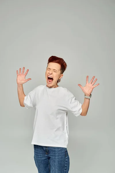 emotional queer person in white t-shirt screaming with closed eyes and gesturing on grey backdrop