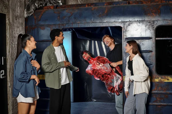 stock image cheerful young man holding creepy mannequin and scaring multicultural friends in escape room