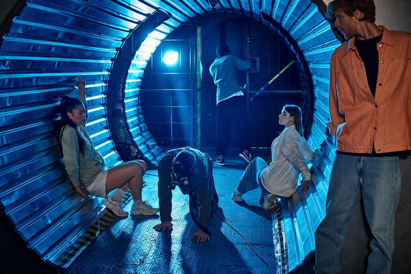 Разнообразная группа друзей, участвующих захватывающее приключение в комнате квестов со страшным человеком в противогазе