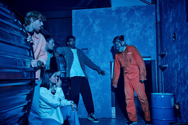 Multicultural group of people hiding from man in gas mask during escape adventure in quest room