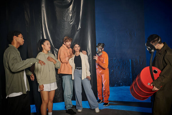 group of scared interracial friends looking at people in ppe suits and gas masks in quest room