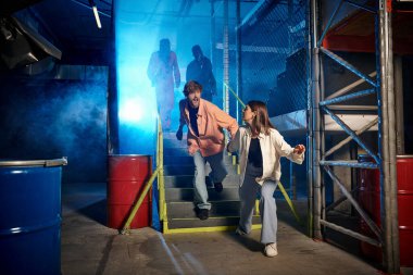 young man and woman running from scary people in gas masks and orange ppe suits in escape room clipart