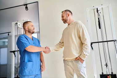 Gülümseyen adam rehabilitasyon merkezinde mavi üniformalı genç doktorla el sıkışıyor.