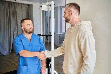 Sakallı adam, rehabilitasyon merkezinde mavi üniformalı gülümseyen bir doktorla el sıkışıyor.