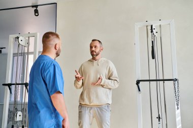 Erkek hasta iyileşme merkezinde mavi üniformalı doktorla tedavi planını tartışıyor.