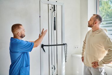Kinesio merkezindeki hastasına egzersiz ekipmanı gösteren mavi üniformalı genç bir doktor.