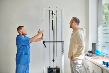 Kinesio merkezindeki hastasına egzersiz ekipmanları gösteren mavi üniformalı bir rehabilitolog.