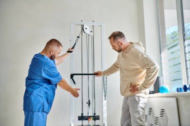 Kurtarma uzmanı mavi üniformalı. Kineziyoloji merkezindeki erkek hastaya eğitim makinesi gösteriyor.