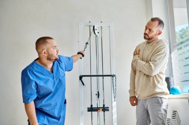 Rehabilitasyon uzmanı Kineziyoloji Merkezi 'nde kolu yaralı bir adama egzersiz ekipmanı gösteriyor.