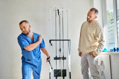 Mavi üniformalı fizyoterapist rehabilitasyon merkezinde eğitim makinesinin yanında talimat veriyor.