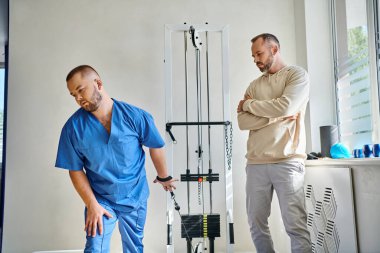 Mavi üniformalı genç doktor rehabilitasyon merkezinde eğitim makinesinin yanında talimat veriyor.