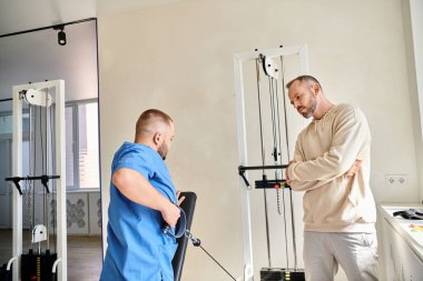 Erkek hasta mavi üniformalı hekime bakıyor. Kineziyoloji merkezinde egzersiz makinesi eğitimi alıyor.