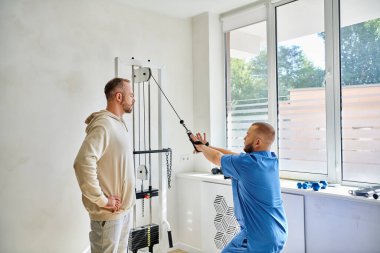 Genç rehabilitasyon uzmanı, erkek hastaya Kinesio merkezinde egzersiz makinesinde nasıl çalışacağını gösteriyor.