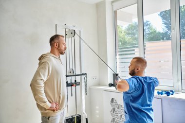 Profesyonel doktor, erkek hastaya Kinesio merkezinde egzersiz makinesinde nasıl çalışacağını gösteriyor.