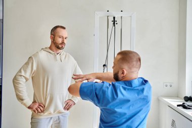 Kinesio merkezindeki hastasına egzersiz makinesini nasıl kullanacağını gösteren mavi üniformalı genç bir doktor.