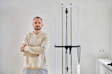 Sakallı adam kollarını kavuşturmuş ve Kinesio merkezinde egzersiz makinesinin yanındaki kameraya bakıyor.