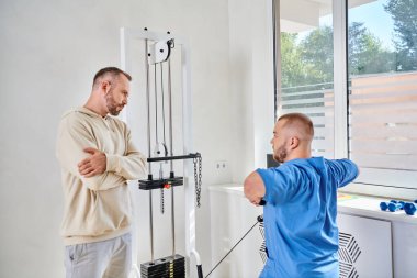 Kolları kavuşturulmuş bir adam rehabilitasyon merkezinde egzersiz makinası eğitimi görüyor.