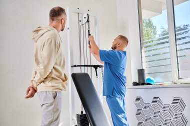 recovery specialist in blue uniform showing training equipment to male patient in kinesio center clipart