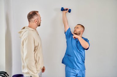 skilled rehabilitologist showing exercise with dumbbell to his patient in recovery kinesio center clipart