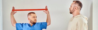 Mavi üniformalı doktor Kinesio merkezinde direniş bandıyla kol egzersizi yapıyor.