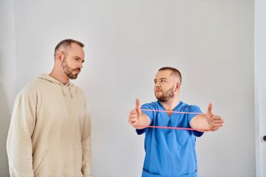 professional recovery specialist showing arm exercise with resistance band to man in kinesio center clipart