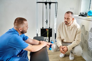 Kinesio Merkezi 'ndeki eğitim makinesinin yanında oturan ve rehabilitasyon uzmanıyla konuşan bir adam.