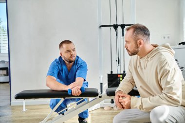 male patient looking at rehabilitologist showing exercise with dumbbell in gym of kinesio center clipart