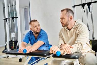 rehabilitologist showing exercise with dumbbell to man in gym of kinesio center, recovery training clipart