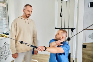 Mavi üniformalı fizyoterapist Kinesio Merkezi 'ndeki adama eğitim makinesinde egzersiz gösteriyor.