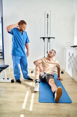 Rehabilitasyon uzmanı Kinesio Merkezi 'nde fitness makinesinin yanında oturan adamla konuşuyor.