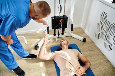 high angle view of instructor assisting his patient lying on fitness man near exercise machine clipart