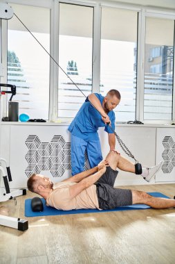 Genç iyileşme uzmanı Kinesio Center spor salonunda egzersiz makinesinde çalışan bir adama yardım ediyor.