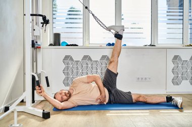 Spor giyimli bir adam Kinesio merkezindeki spor salonunda spor paspasına ve egzersiz makinesine uzanıyor.
