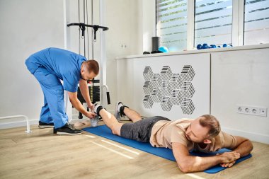 doctor in blue uniform preparing male patient for training on exercise machine in kinesio center clipart