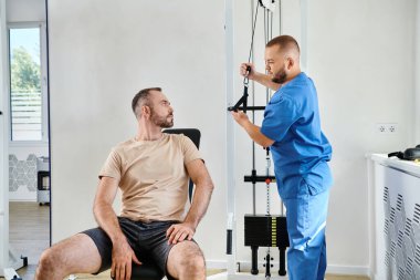 İyileşme uzmanı, erkek hastaya Kinesio merkezinde egzersiz makinesinde nasıl antrenman yapılacağını anlatıyor.