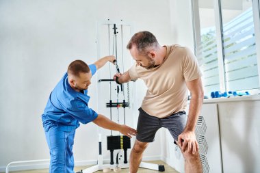 experienced doctor in blue uniform helping man working out on training machine in kinesiology center clipart