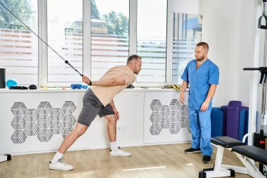 Spor giyimli bir adam Kinesio Merkezi 'ndeki yetenekli rehabilitasyon uzmanının yanında egzersiz makinesinde çalışıyor.