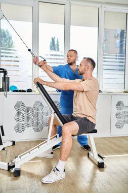 skilled rehabilitologist in blue uniform assisting man during recovery training in kinesio center clipart