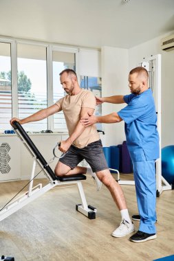experienced specialist supporting man in sportswear during recovery training on exercise machine clipart
