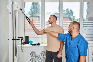 Genç rehabilitasyon uzmanı egzersiz makinesini işaret ediyor ve Kinesio Merkezi 'nin spor salonundaki adama