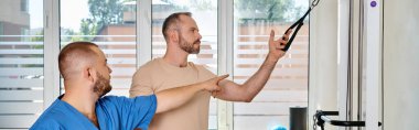 skilled doctor in blue uniform instructing man in gym of kinesio center, horizontal banner clipart