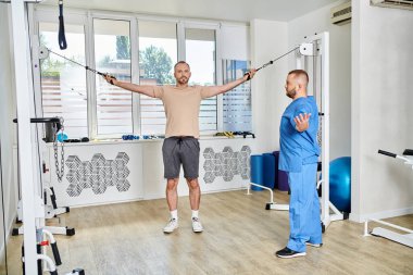 Mavi üniformalı genç doktor Kinesio Center 'da egzersiz makinesinde adama yardım ediyor.