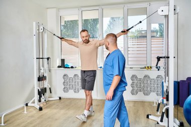 Genç fizyoterapist Kinesio merkezinde egzersiz makinesinde çalışan erkek hastaya yardım ediyor.