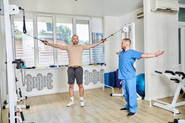 Genç fizyoterapist, Kinesio merkezinde egzersiz makinesinde çalışan erkek hastaya danışmanlık yapıyor.