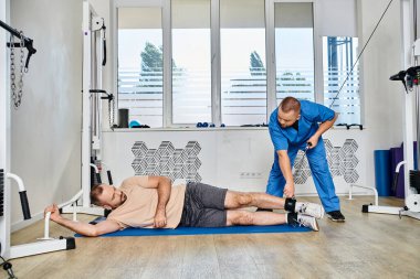 Kinesio Merkezi 'ndeki iyileşme eğitiminde erkek hastaya yardım eden mavi üniformalı genç bir uzman.