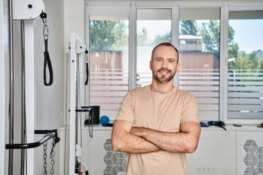 Kollarını kavuşturmuş, modern kineziyoloji merkezinin spor salonunda kameraya bakan mutlu ve yakışıklı bir adam.