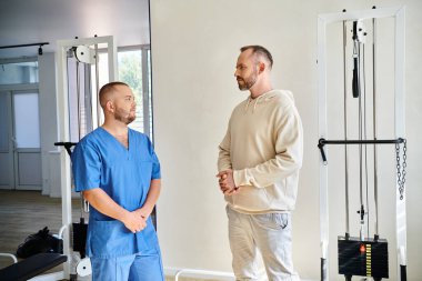 Mavi üniformalı sakallı adam ve profesyonel rehabilitasyon uzmanı modern Kinesio merkezinde konuşuyor.