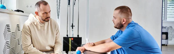 stock image young doctor and man discussing treatment plan near exercise machine in kinesio center, banner